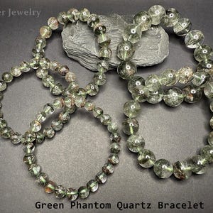 May include: Four green phantom quartz bracelets with clear beads and inclusions. The bracelets are displayed on a dark gray surface. The text "Green Phantom Quartz Bracelet" is at the bottom of the image.