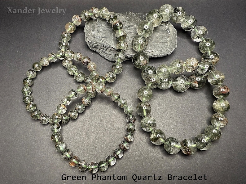 May include: Four green phantom quartz bracelets with clear beads and inclusions. The bracelets are displayed on a dark gray surface. The text "Green Phantom Quartz Bracelet" is at the bottom of the image.
