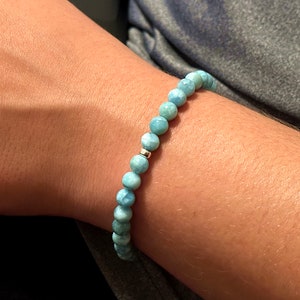 May include: A beaded bracelet featuring round, light blue, marbled Larimar stones. The beads are strung together in a continuous loop, accented by a single, small, polished silver-toned metal bead positioned near the center of the strand.