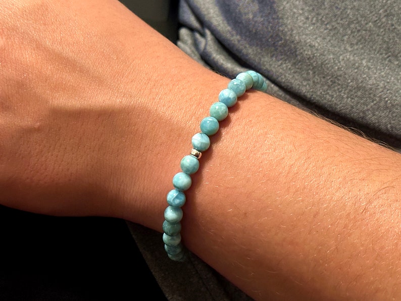 May include: A beaded bracelet featuring round, light blue, marbled Larimar stones. The beads are strung together in a continuous loop, accented by a single, small, polished silver-toned metal bead positioned near the center of the strand.