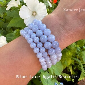 May include: A stack of four blue lace agate bracelets on a person's wrist. The bracelets are made of round beads and are a light blue color. The bracelets are against a background of green leaves and white flowers. The text "Xander Jewelry" and "Blue Lace Agate Bracelet" are visible in the image.