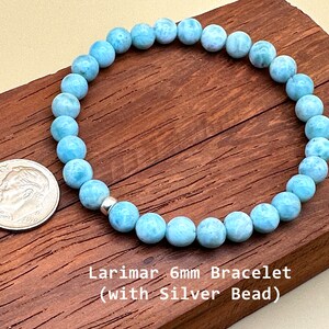 May include: A circular bracelet composed of light blue, marbled Larimar beads measuring 6mm in diameter, featuring a single metallic silver bead. A US dime is placed next to the bracelet for scale, and the text reads Larimar 6mm Bracelet.