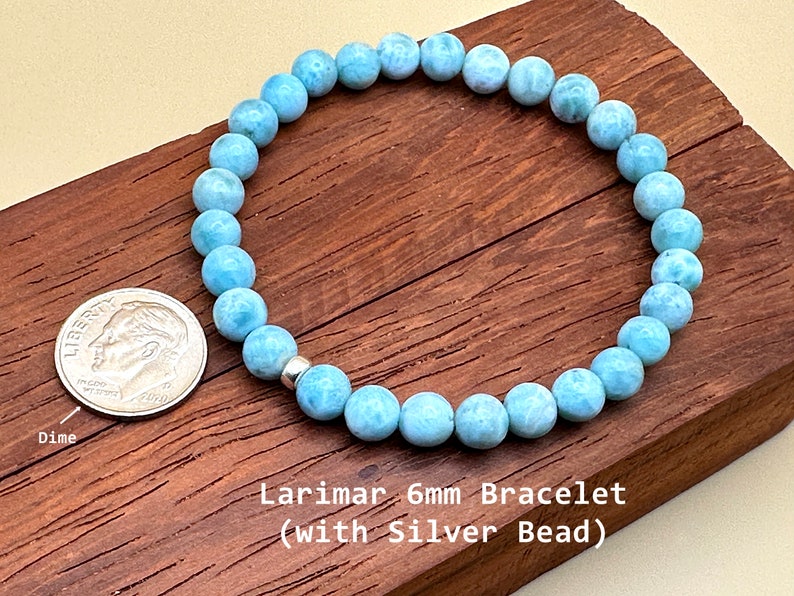 May include: A circular bracelet composed of light blue, marbled Larimar beads measuring 6mm in diameter, featuring a single metallic silver bead. A US dime is placed next to the bracelet for scale, and the text reads Larimar 6mm Bracelet.