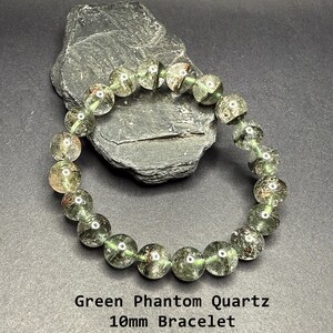 May include: Green Phantom Quartz bracelet with 10mm beads. The bracelet features translucent green beads with inclusions, strung together on an elastic band. The bracelet is displayed on a dark gray surface with a dark gray rock in the background. The text "Green Phantom Quartz 10mm Bracelet" is at the bottom.