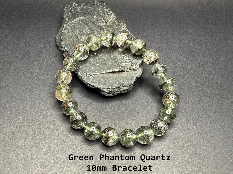 May include: Green Phantom Quartz bracelet with 10mm beads. The bracelet features translucent green beads with inclusions, strung together on an elastic band. The bracelet is displayed on a dark gray surface with a dark gray rock in the background. The text "Green Phantom Quartz 10mm Bracelet" is at the bottom.