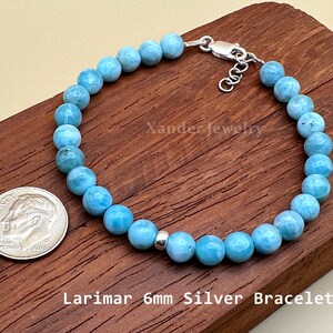 May include: A bracelet featuring light blue, round Larimar beads, a single silver spacer bead, and a silver lobster clasp. The text Xander Jewelry and Larimar 6mm Silver Bracelet are visible. A US dime is included for scale next to the jewelry piece.