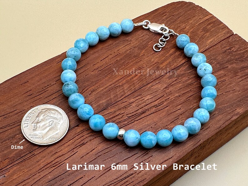 May include: A bracelet featuring light blue, round Larimar beads, a single silver spacer bead, and a silver lobster clasp. The text Xander Jewelry and Larimar 6mm Silver Bracelet are visible. A US dime is included for scale next to the jewelry piece.