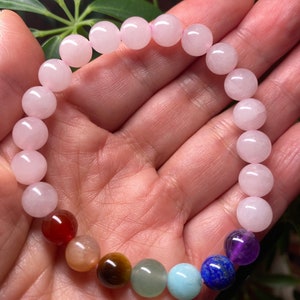 May include: A beaded bracelet featuring a series of smooth, round, pale pink rose quartz beads combined with a single set of seven multi-colored gemstone beads in shades of deep red, tan, brown, green, light blue, dark blue, and purple.