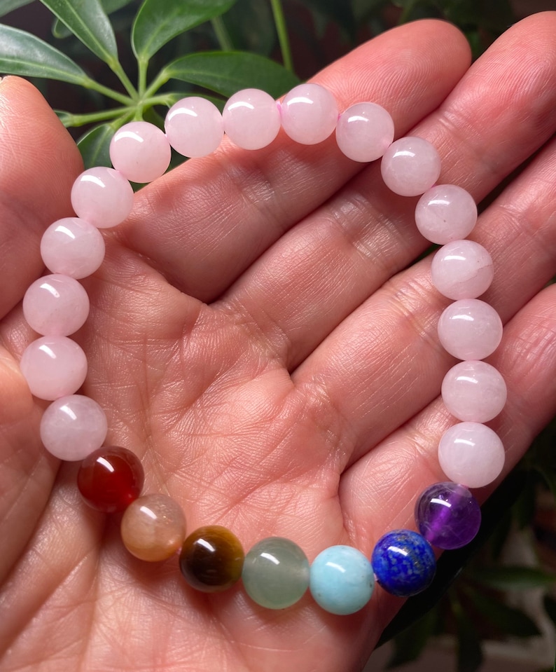 May include: A beaded bracelet featuring a series of smooth, round, pale pink rose quartz beads combined with a single set of seven multi-colored gemstone beads in shades of deep red, tan, brown, green, light blue, dark blue, and purple.