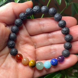 May include: A beaded bracelet featuring a series of porous, black lava stone beads and seven smooth, polished gemstone beads in a spectrum of colors including red, orange, yellow, green, light blue, dark blue, and purple, arranged in a circular row.