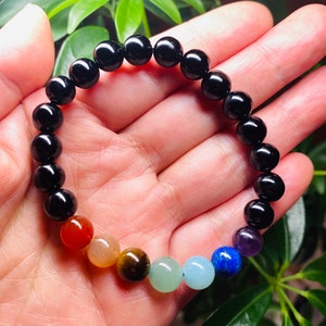 May include: A beaded bracelet featuring a series of smooth, round, polished black beads interspersed with a sequence of seven distinct colored stones including red, orange, brown, light green, light blue, dark blue, and purple spherical beads.