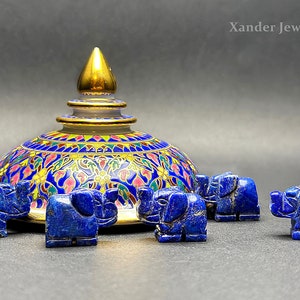 Puede incluir: Cinco pequeñas figuritas de elefante de lapislázuli azul rodean una cúpula de cerámica decorativa con intrincados patrones florales en tonos azules, rosas y dorados. La cúpula tiene un remate dorado en la parte superior.  Xander Jewelry está impreso en el fondo.