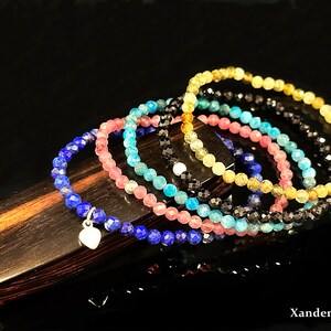 May include: A set of six gemstone bracelets, each featuring a different color: blue, turquoise, pink, black, and two shades of yellow. The bracelets are stacked on top of each other, with a silver heart charm hanging from the blue bracelet. The bracelets are displayed on a wooden surface.