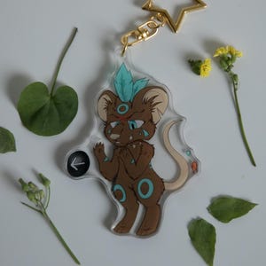 May include: A clear acrylic keychain featuring a cartoon mouse character with brown fur, teal accents, and a teal feather. The keychain has a gold star-shaped clasp. The image is decorated with leaves and small yellow flowers.