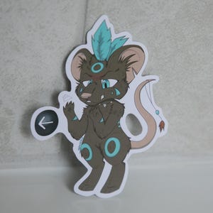 May include: A sticker featuring a cartoon mouse-like creature with brown fur, teal accents, and a teal feather. The mouse has a stern expression and is holding a sign with a left-pointing arrow. The sticker is on a white background.