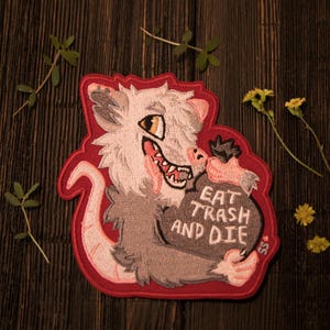 May include: Embroidered patch with a cartoon opossum design and a red border. The opossum is depicted in grey and white, holding a bag with the text "EAT TRASH AND DIE". The patch is set against a dark wooden background.
