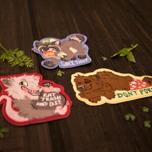 May include: Three embroidered patches with cartoon animal designs. One shows a raccoon labelled "Snack Thief", another a wolf with "Eat Trash and Die", and a bear with "Don't Poke". The patches are on a dark wood surface.