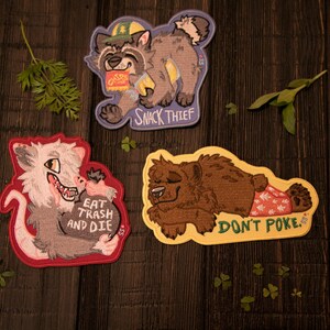 May include: Three embroidered patches with animal illustrations on a dark wood surface. One patch features a raccoon with the text "SNACK THIEF." Another shows an opossum with the text "EAT TRASH AND DIE." The third depicts a bear with the text "DON'T POKE."