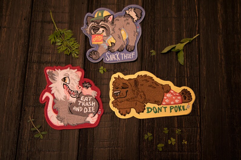May include: Three embroidered patches with animal illustrations on a dark wood surface. One patch features a raccoon with the text "SNACK THIEF." Another shows an opossum with the text "EAT TRASH AND DIE." The third depicts a bear with the text "DON'T POKE."