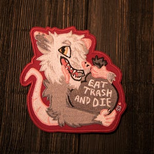 May include: Embroidered patch depicting a cartoon opossum with a wide grin, holding a bag with the text "EAT TRASH AND DIE." The patch has a red border and is set against a dark wood grain background.