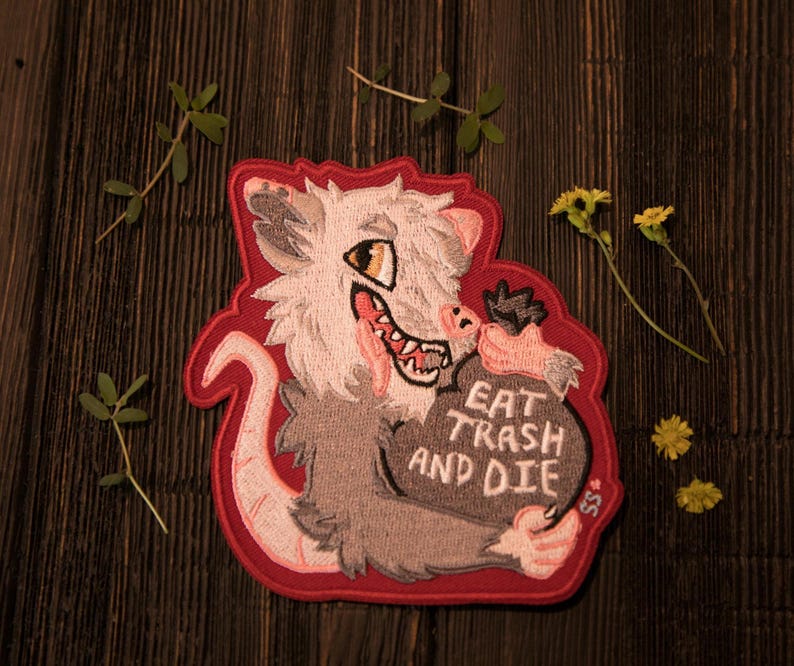 May include: Embroidered patch with a cartoon opossum design and a red border. The opossum is depicted in grey and white, holding a bag with the text "EAT TRASH AND DIE". The patch is set against a dark wooden background.