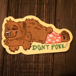 May include: Embroidered patch showing a cartoon bear in a relaxed pose, wearing patterned shorts. The patch has the text "DON'T POKE" in green. The bear is brown with a yellow border, suitable for jackets, bags, or other fabric items.