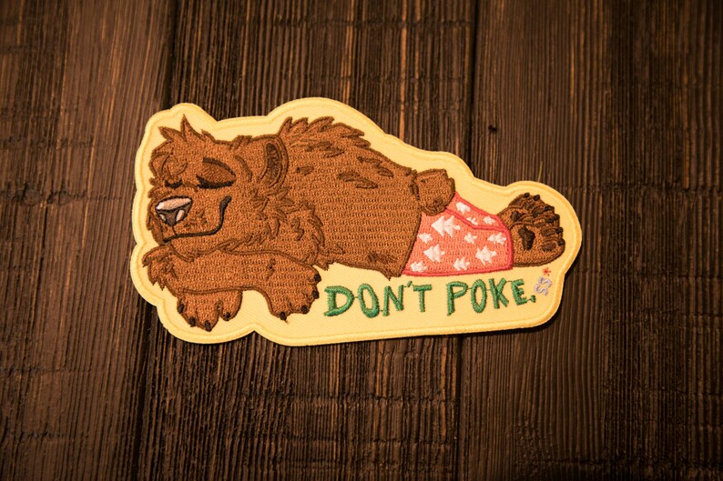May include: Embroidered patch showing a cartoon bear in a relaxed pose, wearing patterned shorts. The patch has the text "DON'T POKE" in green. The bear is brown with a yellow border, suitable for jackets, bags, or other fabric items.