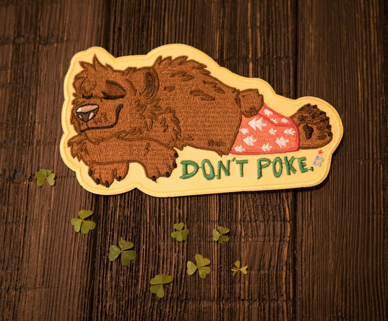 May include: Embroidered patch showing a brown bear in pyjamas, lying down with closed eyes. The patch has a yellow border and the text "DON'T POKE" in green. The bear's pyjamas are red with white star patterns.
