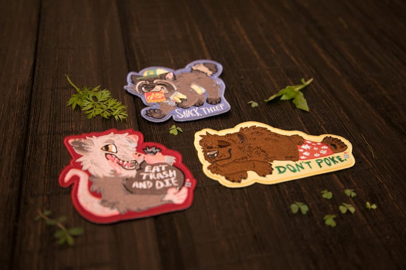 May include: Three embroidered patches with cartoon animal designs. One shows a raccoon labelled "Snack Thief", another a wolf with "Eat Trash and Die", and a bear with "Don't Poke". The patches are on a dark wood surface.
