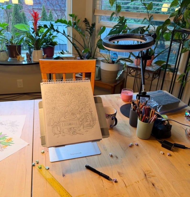 Peut inclure: Une table en bois avec un livre de coloriage sur un support, une lampe annulaire et diverses fournitures artistiques. Le livre de coloriage est ouvert sur une illustration d&eacute;taill&eacute;e. Des crayons de couleur, une tasse et un stylo sont &eacute;galement visibles.