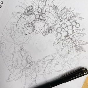 K&ouml;nnte beinhalten: Eine Bleistiftskizze eines Kranzdesigns mit Erdbeeren, Blaubeeren und Bl&auml;ttern. Die Zeichnung ist detailliert mit Schattierungen und Umrissen, und ein schwarzer Stift befindet sich im Vordergrund.