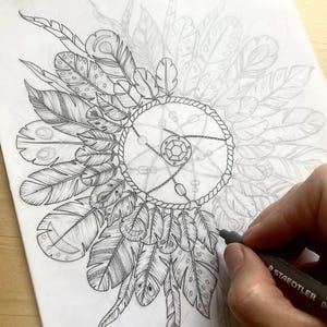 K&ouml;nnte beinhalten: Eine detaillierte Bleistiftskizze eines Traumf&auml;ngers mit Federn. Der zentrale Kreis ist mit einem geometrischen Design und h&auml;ngenden Ketten verziert. Die Hand des K&uuml;nstlers ist sichtbar, die einen schwarzen Marker h&auml;lt und der Zeichnung Details hinzuf&uuml;gt.