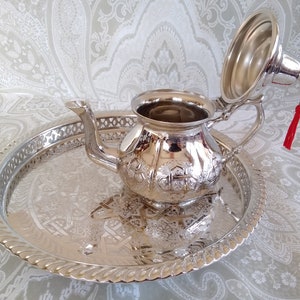 May include: A silver Moroccan teapot with an ornate design sits on a silver tray with a matching design. The teapot has a lid with a tassel.