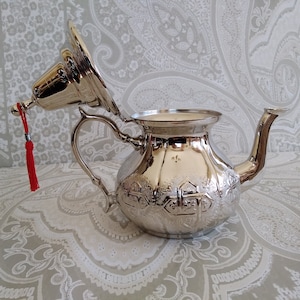 May include: A silver teapot with an ornate design and a red tassel. The lid is open and the teapot is sitting on a patterned surface.