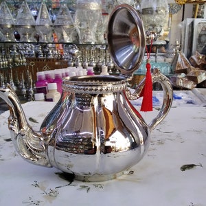 May include: A silver teapot with a shiny, polished finish and a red tassel on the lid. The teapot has a decorative spout and handle.