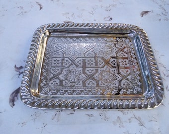 Peut inclure: Un plateau de service rectangulaire argenté avec un motif floral et géométrique orné au centre. Le plateau a un motif de corde torsadée sur les bords. Le plateau est probablement en métal et convient pour servir des aliments ou exposer des objets.
