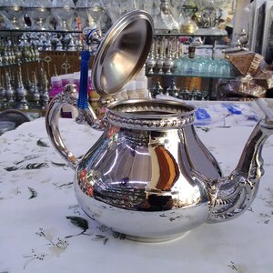 May include: A silver teapot with a decorative handle and spout. The lid is open and has a blue tassel. The teapot is sitting on a white tablecloth with a floral pattern.