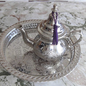 May include: A silver teapot with an ornate design and a purple tassel sits on a silver tray with an intricate pattern.