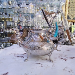 May include: A silver teapot with intricate engraved designs and a turquoise tassel. The teapot has a round body and a lid that is open. The teapot is sitting on a white tablecloth with a floral pattern.