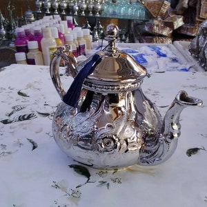 May include: A shiny silver teapot with intricate floral and scrollwork designs. The teapot has a curved spout, a handle, and a lid with a small knob. A dark blue tassel hangs from the lid. The teapot sits on a white floral patterned tablecloth.