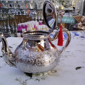 May include: A silver teapot with an ornate design and a red tassel. The teapot has a hinged lid and a spout. The teapot is sitting on a white tablecloth.