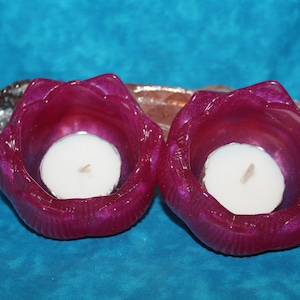 May include: Two magenta-colored, flower-shaped candle holders with white candles inside. The candle holders are on a blue background. A piece of crystal is behind the candle holders.