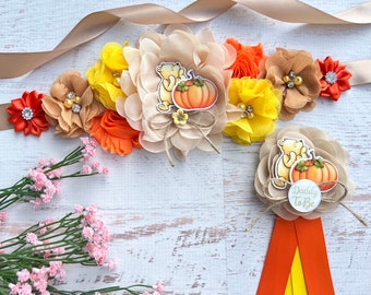 May include: A brown sash with a large flower arrangement in shades of orange, yellow, and brown. The flowers are made of fabric and have a rustic, fall theme. The sash is decorated with a Winnie the Pooh character holding a pumpkin. A smaller version of the flower arrangement is attached to a yellow and orange ribbon with the text "Daddy to Be".