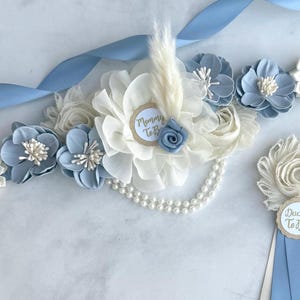 K&ouml;nnte beinhalten: Ein Babyparty-Set mit Stoffblumen in Wei&szlig; und Hellblau. Das Set besteht aus einer Sch&auml;rpe mit hellblauem Band und einer Perlenkette. Der Anstecker hat einen "Daddy To Be" Anh&auml;nger. Die Sch&auml;rpe hat einen "Mommy To Be" Anh&auml;nger.