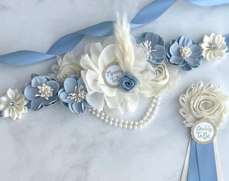 K&ouml;nnte beinhalten: Ein Babyparty-Set mit Stoffblumen in Wei&szlig; und Hellblau. Das Set besteht aus einer Sch&auml;rpe mit hellblauem Band und einer Perlenkette. Der Anstecker hat einen "Daddy To Be" Anh&auml;nger. Die Sch&auml;rpe hat einen "Mommy To Be" Anh&auml;nger.