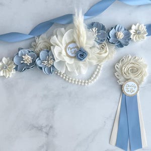 K&ouml;nnte beinhalten: Hellblaues und wei&szlig;es Blumen-Set f&uuml;r eine Babyparty, bestehend aus einer Sch&auml;rpe und einer Anstecknadel. Die Sch&auml;rpe hat Stoffblumen, Perlen und ein Band. Die Anstecknadel tr&auml;gt die Aufschriften "Daddy To Be" und "Mommy To Be".