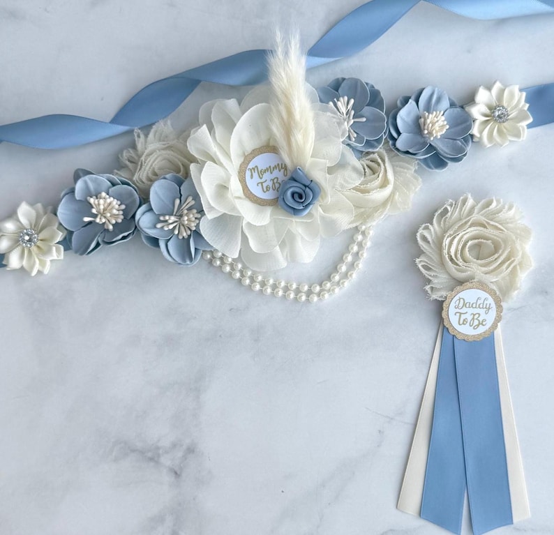 K&ouml;nnte beinhalten: Hellblaues und wei&szlig;es Blumen-Set f&uuml;r eine Babyparty, bestehend aus einer Sch&auml;rpe und einer Anstecknadel. Die Sch&auml;rpe hat Stoffblumen, Perlen und ein Band. Die Anstecknadel tr&auml;gt die Aufschriften "Daddy To Be" und "Mommy To Be".