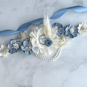 K&ouml;nnte beinhalten: Ein hellblaues Satinband, verziert mit Stoffblumen in Wei&szlig;- und Blaut&ouml;nen. Ein zentrales Arrangement zeigt ein Etikett mit der Aufschrift "Mommy To Be", eine kleine blaue Rose und eine Feder. Eine Perlenkette und weitere Blumen vervollst&auml;ndigen das Design.