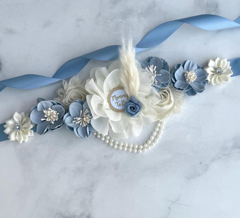 K&ouml;nnte beinhalten: Ein hellblaues Satinband, verziert mit Stoffblumen in Wei&szlig;- und Blaut&ouml;nen. Ein zentrales Arrangement zeigt ein Etikett mit der Aufschrift "Mommy To Be", eine kleine blaue Rose und eine Feder. Eine Perlenkette und weitere Blumen vervollst&auml;ndigen das Design.