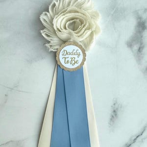 K&ouml;nnte beinhalten: Eine wei&szlig;e Stoffblume mit einem goldumrandeten runden Anh&auml;nger mit der Aufschrift "Daddy To Be". Der Anh&auml;nger ist an einem Band mit einem hellblauen Streifen und einem cremefarbenen Streifen befestigt, f&uuml;r eine Babyparty oder Gender Reveal.